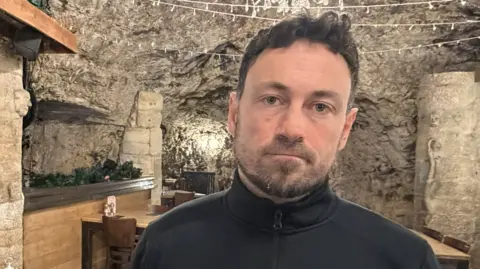 Phil Steel is standing in the restaurant, which is built into a cave, and is wearing a grim expression. He has short black hair and a beard.