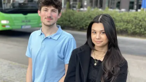 A man with brown hair in a blue polo shirt and a girl with black hair in a black coat and top sit outside with a green bus in the background. They are smiling at the camera