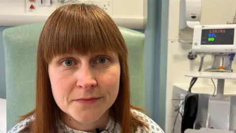 A woman in her mid 30s is sitting in a hospital room, on a green chair and is wearing a gown. She has been prepped for theatre. She has red hair that is shoulder length with a full blunt fringe. 