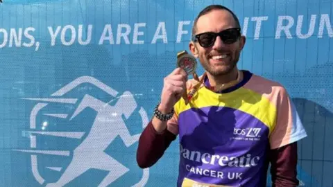 Peter Dunlop A man with brown hair and a brown beard holding a marathon medal and smiling in a Pancreatic Cancer UK t-shirt.