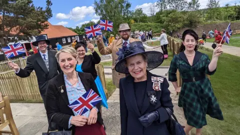 Beamish Museum Sue snowdon sporting a magnificent hat with Beamish staff members dressed in 1950s clothing waving flags