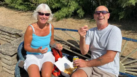 A couple sit on a bench smiling. Penny is wearing a vest top and shorts, John is wearing a grey T-shirt and both are wearing sunglasses. They have some chips in a cardboard carton and John is holding a fizzy drink in a can.