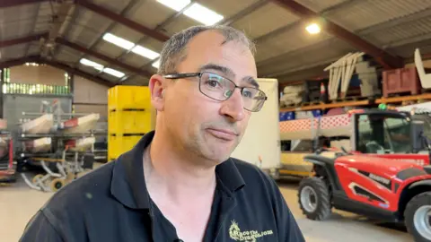Image shows Dan Windsor, Operations Director at Race the Dragon. He is wearing a black polo shirt which says 'Race The Dragon' in gold. He has short dark hair and is wearing glasses. He is looking off to the right. Behind him to the left is dragon boats. To his right, is a fork truck which is bright red.