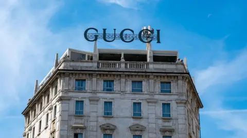 A Gucci logo on the roof of a stone building in Milan, beneath a bright blue sky.