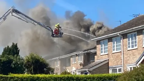 Firefighters on an aerial platform spray water on to a smoldering roof of a row of terraced housing. 