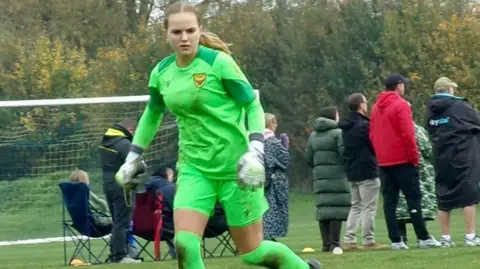 Amelia Aplin has long blonde hair and is wearing a bright green goalkeeper kit with white gloves.