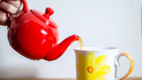  English breakfast tea being poured from a red teapot into a mug with a yellow flower design on it.