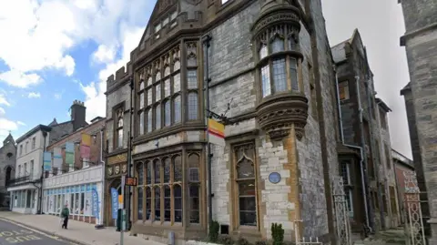 Google The front of the Dorset Museum on the sloping hill of High West Street - a stone building with large gothic-style bay windows with leaded glass on the ground and first floor and a crenellated parapet with a pitched roof