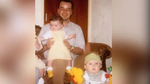 Folheto para a família Joseph está sentado em um sofá com a filha no colo. Dan Astles, que tem apenas alguns meses de idade, está sentado em uma espreguiçadeira colorida à sua frente nesta fotografia.