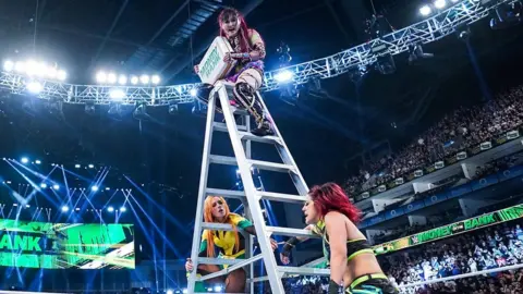 WWE Wrestler Iyo Sky perches on top of a stepladder in the middle of the ring during the women's Money in the Bank match. She's clutching a briefcase, with Money in the Bank written on it. Becky Lynch is handcuffed to the bottom of the ladder at the left while Bayley is handcuffed to the opposite side.