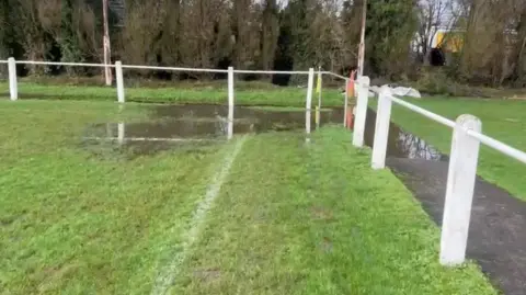 Paul Tudor A grass football pitch with large areas of standing water pooling across the surface, with white perimeter railings and bare winter trees in the background.