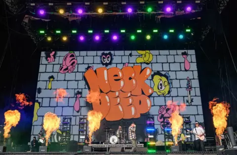 Getty Images The five members of pop-punk band Neck Deep perform on the main stage at Leeds Festival. Fire comes out of the stage whilst they are stood in front of a colourful screen reading the name of the band.