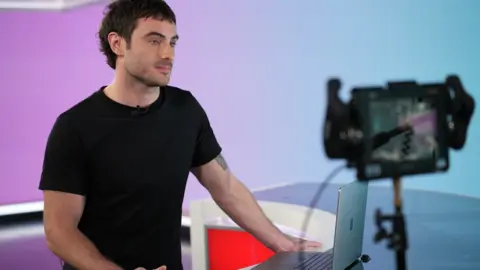 Presenter is standing on at a desk on a television set, he is typing on a laptop, you can see a TV camera recording him