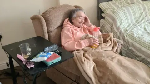 A  woman on a sofa with her feet up looking frail. She is drinking from a sports cup and there are some items such as a hairbrush and baby wipes on a table next to her. 