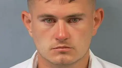 A police "mugshot" photo of Stephen Rennie. He is clean shaven, has dark hair and green eyes. He is wearing a white shirt.