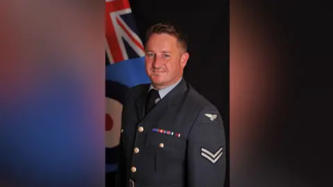 RAF veteran David Morris is dressed in a navy blue military uniform. He smiles as he looks at the camera. A Union Jack flag can be seen in the background of the photo. 