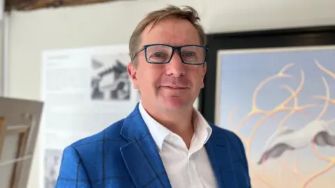 Grant Ford in a blue blazer and white shirt in front of a surrealist painting. He wears glasses as he looks into the camera.