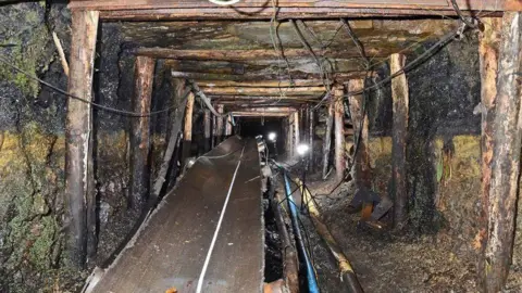 Crown Prosecution Service Inside the Gleision drift mine, with wires and a conveyor belt inside a rock walls held up by wooden beams going deep underground.