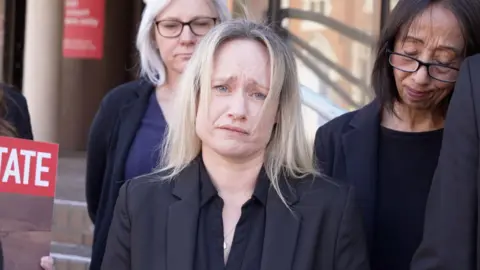 A woman with blonde hair wearing all black. She looks devastated and is holding back tears. There are people standing behind her also looking very upset.