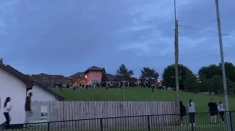 A number of young people can be seen congregating on a grassy embankment near a large pink building. Telephone wires and a picket fence can be seen at the bottom of the hill close to a white building.