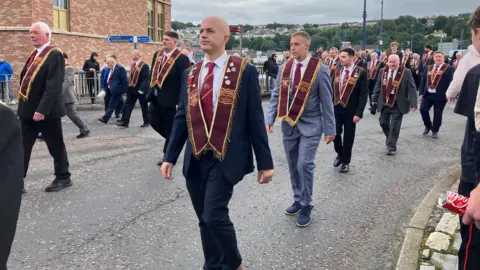 A group of men wearing black and grey suits marching in a parade. They are wearing maroon sashes an carrying various flags.