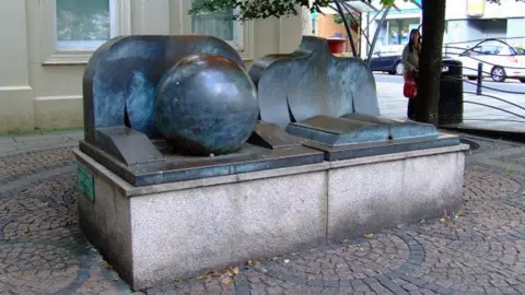 A large sculpture on a street, showing an open book, a large globe and two chests and arms behind them. 