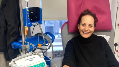 CRUK Donna smiles at the camera as she's linked to an oncology machine which is providing her treatment. She is wearing a black turtle neck jumper. Her hair is tied back into a ponytail and she has her glasses on her forehead.