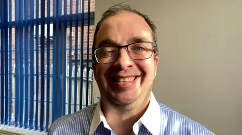 A man with short, thinning dark hair and glasses smiles at the camera. He is standing indoors, in front of a window and wearing a blue and white striped shirt.