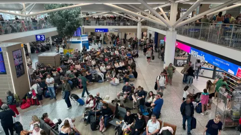 BBC Inside the departure lounge at Bristol Airport. There is a large waiting area with chairs in the middle of the airport and shops at the side. Many passengers are sat down waiting to depart, while others are walking around.
