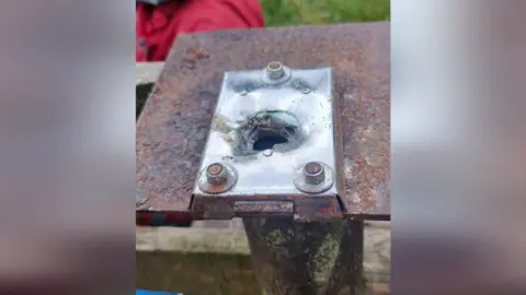 The vandalised box. It shows rusty metal sheet with a hinged plate that has been perforated.