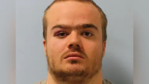 Met Police A mugshot of Jonty Bravery, who is wearing a grey shirt. He is staring at the camera and has a beard and short, blonde hair.