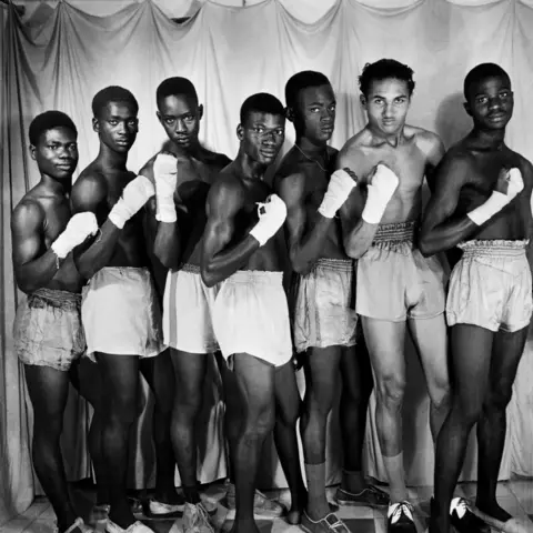 Roger DaSilva/Josef and Anni Albers Foundation Men dressed in boxing shorts with bandaged hands pose in a line for the camera.