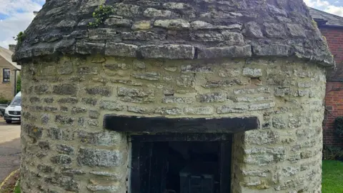 David Brough A close up of a stone-built round house with a small black door. The stonework on the bottom has been cleaned and restored and looks in good condition. The stonework on the roof is dark in colour and has greenery growing between the bricks. 
