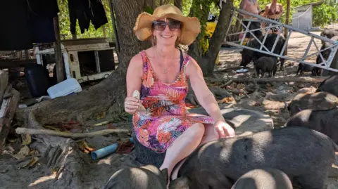 Supplied A woman in a palm leaf print, red, white and black sun dress sits on an old tyre while stroking one of a group of black pigs snuffling in the ground. She is smiling at the camera and is wearing sunglasses and a straw hat. 