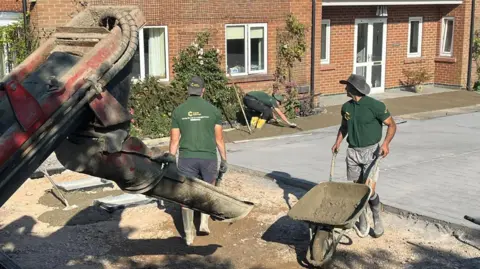 Bradley Smith Two men in khaki green t-shirts with yellow lettering are wearing hats and carrying wheelbarrows. A red digger is at the front on the photo. They are in the process of digging up and block paving a driveway. 