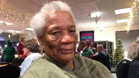 Kate Bradbrook/BBC Woman with white hair wearing a green top in a room full of people