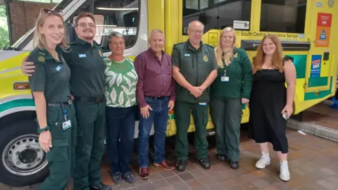 SCAS Barry Crawford, a man in blue jeans and a red shirt, and his wife Alison, wearing a green top and jeans, stand with four paramedics in front of an ambulance.