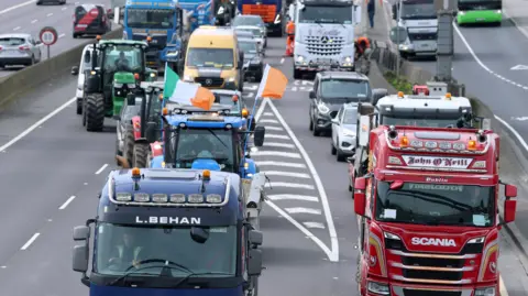 Lorries and tractors are on a busy road. One tractor has Irish tricolour flags.