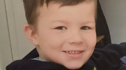Family image caio, victim of a dog attack, smiling on the camera. The young boy has short brown hair and brown eyes and wears the dark bird.