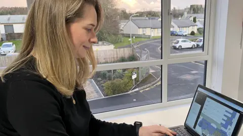 Rosie Richardson sitting at a desk looking at her laptop with the tracking technology shown on the screen. She has shoulder-length blonde hair. She is wearing a black jumper. She is in front of a window at sunset. It looks out to a housing estate with Cockermouth Castle visible in the skyline.