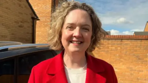A woman smiles at the camera. She has short blonde hair and is wearing a red jacket and white t-shirt. 