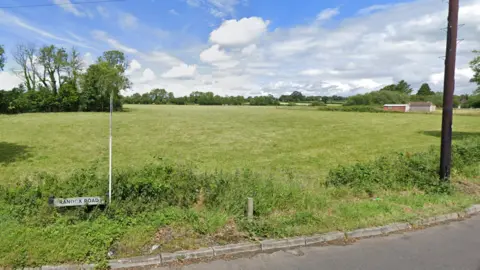 A green field above a sign for Randox Road. It's a sunny day with blue skies and a few clouds. 