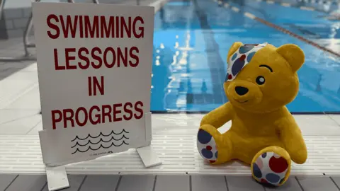 BBC Children in Need mascot Pudsey the Bear sits on the edge of an indoor swimming pool, next to a sign that says "Swimming lessons in progress" in large red letters. Pudsey is bright yellow, with polka dots on his ears, paws, and a patch over one eye