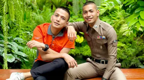 Pisit Sirihirunchai Pisit in his police unform sitting next to Chanatip with his arm on Chanatip's shoulder. Behind them is a lush garden. 