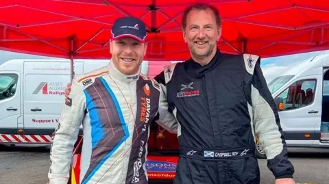Sir Chris Hoy and Campbell Roy stand arm in arm, wearing drivers outfits with two vans in the background. They are standing under a canopy.