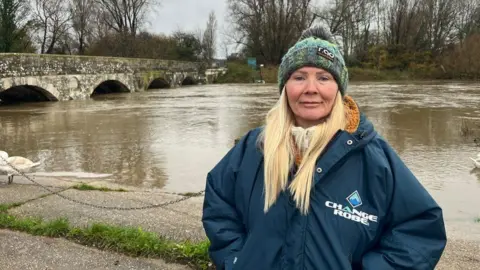 Woman with long very light blonde hair wearing a hat and coat - a river and bridge are behind her.