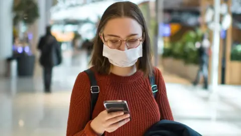 Getty Images Stock image of a young woman wearing a face mask and holding a smartphone