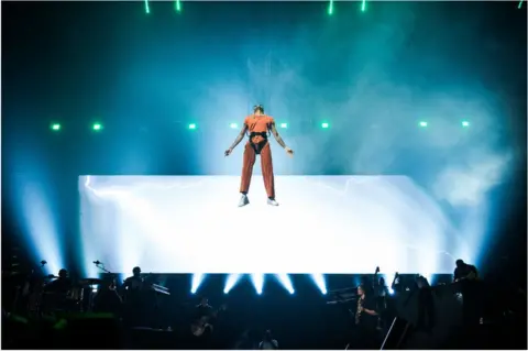 Getty Images Wizkid suspended in the air, wearing a yellow jumpsuit. There are brigh light behind him.