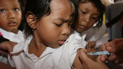 AFP Girl watches as an adult hand administers a vaccine by needle into her upper arm. Child next to her also watches