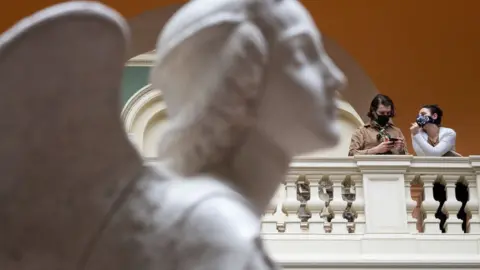 Leon Neal/Getty Images Two women wear masks as they chat with a marble statue in the foreground